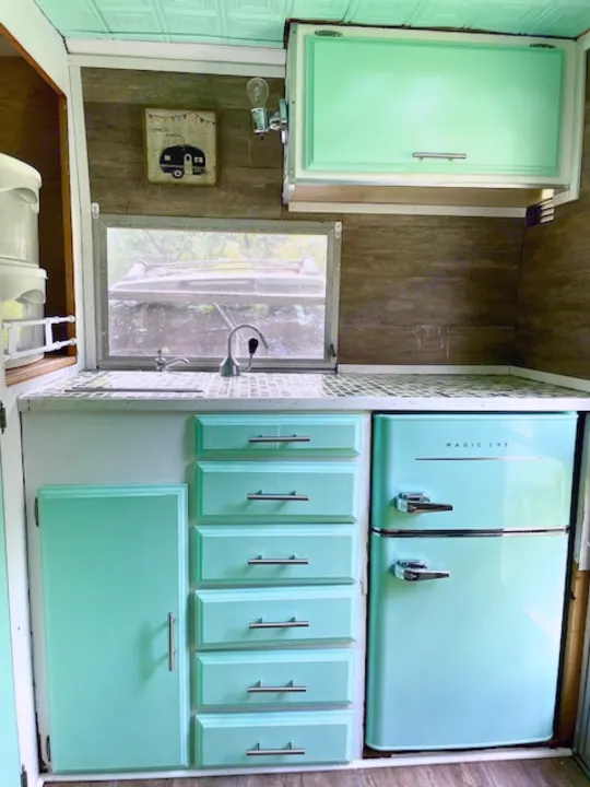 Inside a retro-themed RV rental kitchen, featuring mint green cabinets, a compact fridge, and a small sink under a window.. Shasta Compact 1973