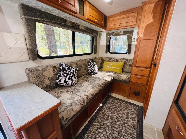Cozy RV rental dinette with patterned cushions, overhead wood cabinets, and windows revealing a scenic lake view.. Bigfoot RV Bigfoot RV Trailer 2005