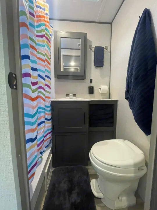 Compact RV rental bathroom featuring a colorful striped shower curtain, toilet, vanity, and ample storage.. Dutchmen Aspen Trail 2022