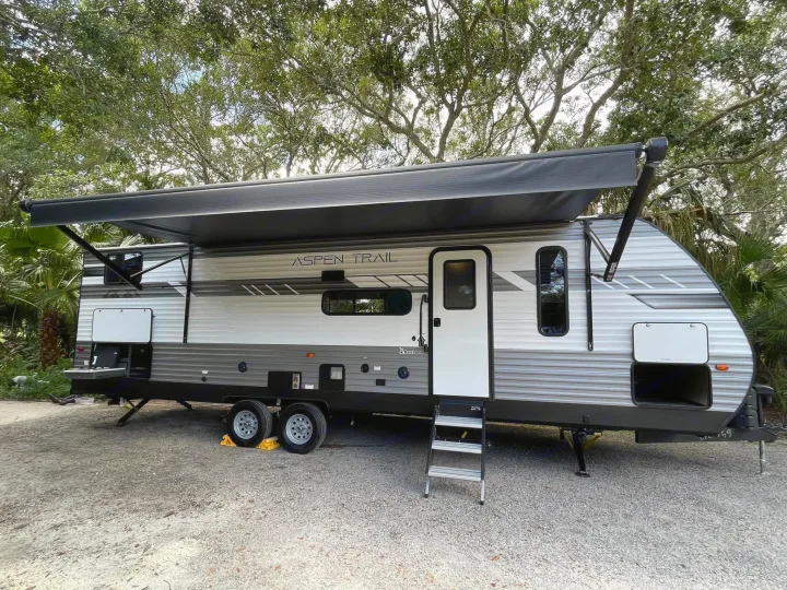Gray and white Aspen Trail RV rental parked on a gravel campsite. Awning extended, steps down, and storage open.. Dutchmen Aspen Trail 2022