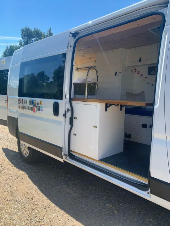 White RV rental conversion van with open side door revealing a custom interior, including a kitchen and bed area, for van life.. Dodge Promaster 2500 2021