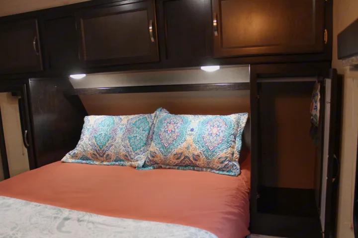 Comfortable RV rental bedroom with a bed, colorful patterned pillows, peach sheets, dark wood cabinets, lights & storage.. Coachmen Spirit Ultralight 2020