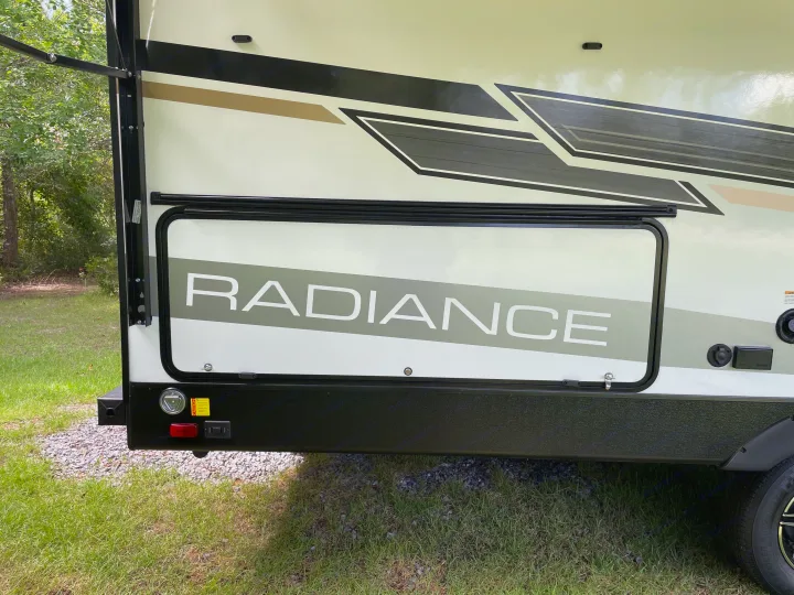 Side view of an RV rental with "RADIANCE" text, black and tan graphics, and a storage door, parked on grass.. Cruiser Rv Corp Radiance 2022