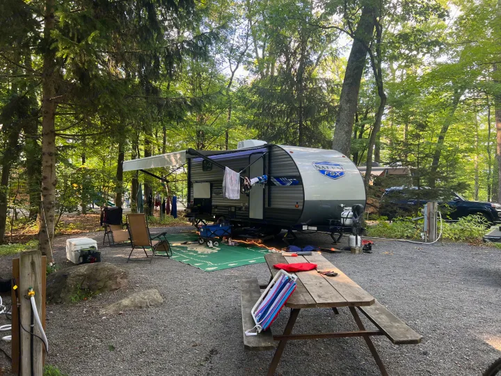 An RV rental is set up at a peaceful, gravel campsite, surrounded by tall trees with a picnic table and gear.. Forest River Salem FSX 2022