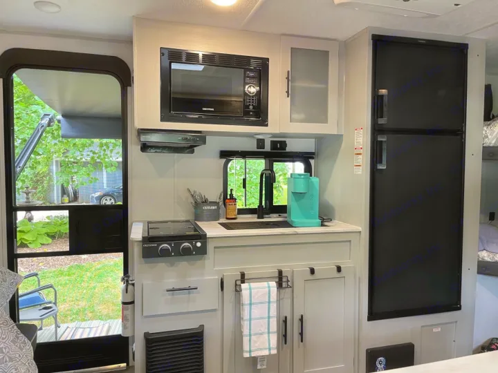 Compact RV rental kitchen featuring a microwave, sink, and fridge, with an open door view to a green yard. Great for camping.. Forest River Wildwood FSX Plantium 2022