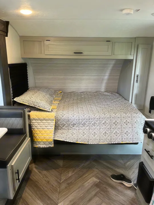 RV rental bedroom with patterned grey/yellow bed, light wood cabinets, and herringbone flooring. Dinette to left.. Forest River Wildwood FSX Plantium 2022