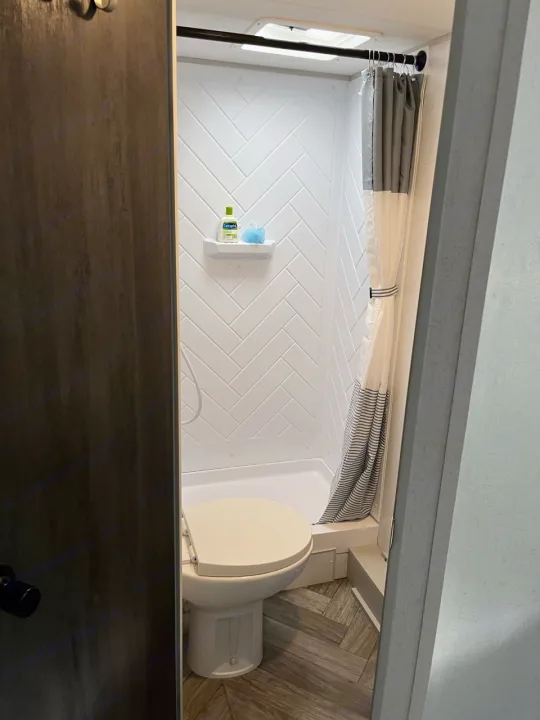 RV rental bathroom features: compact shower with white herringbone walls, toilet, and wood-look flooring.. Forest River Wildwood FSX Plantium 2022