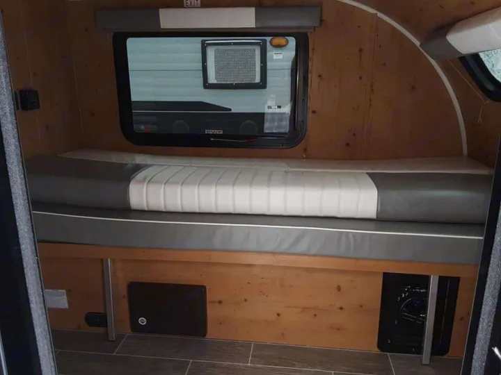Inside a comfy rv rental: grey and white cushioned bench seating/bed, wood walls, and a large window.. Sunset Park RV SunRay 2021