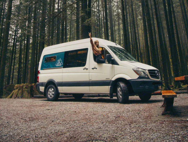Ready for adventure! A woman gives a peace sign from a white Sprinter **RV rental** van parked in a lush forest campsite.. Mercedes Sprinter 2016