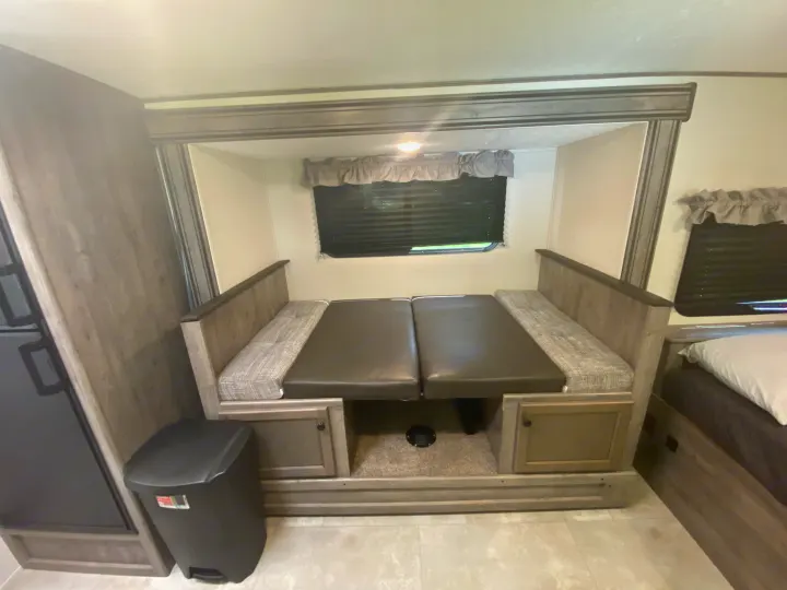 Inside an RV rental: a dinette area converted into a bed with gray and black cushions, under-seat storage, window, and trash can.. Coachmen Apex Nano 2021