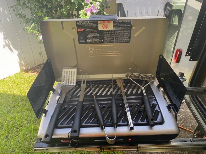 Coleman portable grill with BBQ tools, set up for outdoor cooking next to an RV rental. Perfect for camping meals.. Coachmen Apex Nano 2021