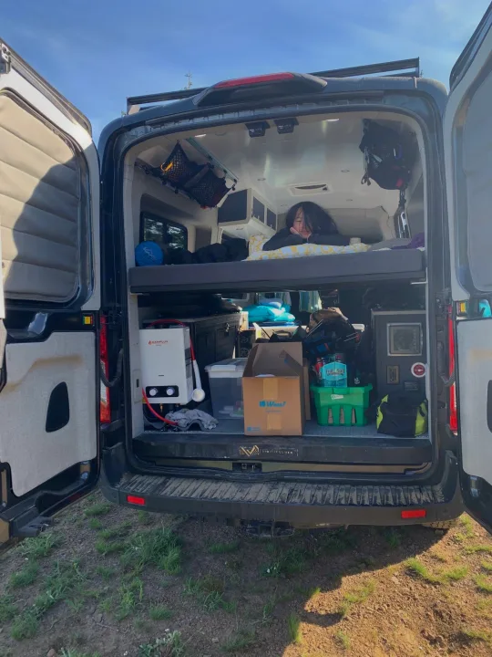 Inside an **RV rental**, a person rests on the top bunk of a camper van. Below, organized storage & portable shower for van life.. Other Transit 2020