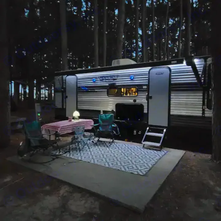 Nighttime RV rental campsite scene with illuminated camper, picnic table, chairs, and outdoor rug under tall trees.. Forest River Salem Cruise Lite 2022