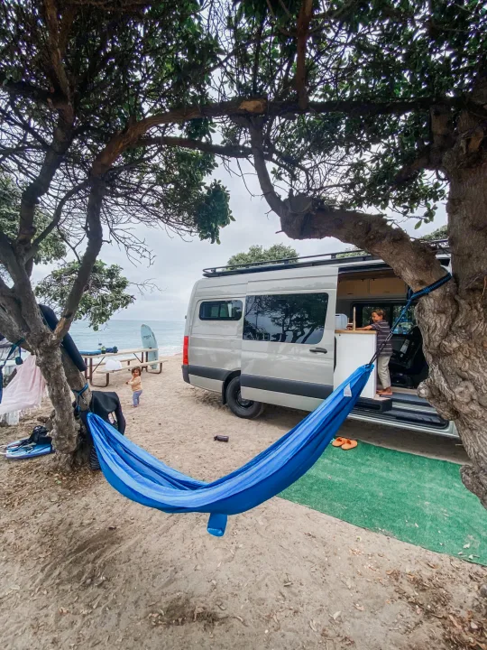An RV rental provides beach camping comfort. Family enjoys the ocean view with a blue hammock strung between trees and the van.. Mercedes Sprinter 2021