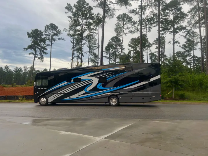 This black Outlaw RV rental, with blue & white graphics, is parked on wet pavement near tall pine trees, ready for adventure.. Thor Outlaw 2022