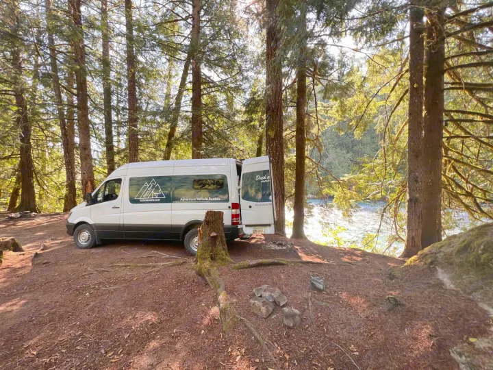 White camper van (RV rental) nestled in a sunlit forest clearing beside a river, near a fire pit.. Mercedes Sprinter 2016