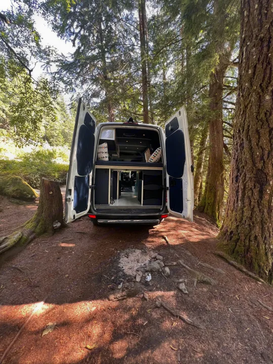 Rear view of an RV rental camper van parked in a forest campsite, doors open to a cozy interior, surrounded by tall trees and a dirt path.. Mercedes Sprinter 2016