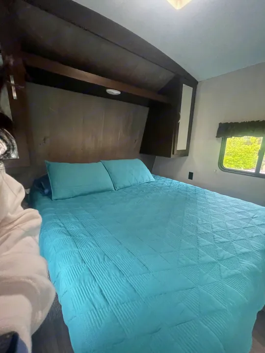 Inside a comfortable RV rental bedroom, featuring a blue bed with pillows, overhead cabinets, and a window view.. Keystone RV Springdale 2020