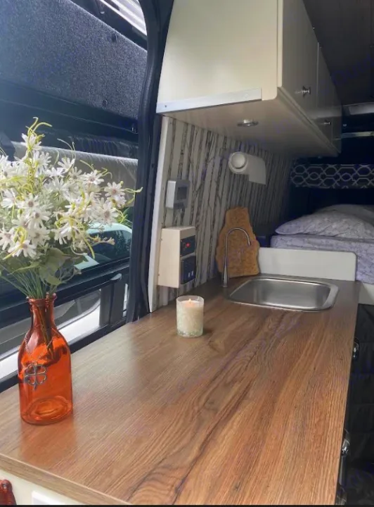 Cozy RV rental camper interior: wooden counter with sink & faucet, white cabinets, and bed. Flowers & candle add charm.. Mercedes Sprinter 2012