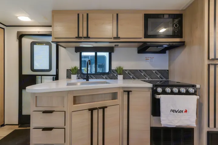Compact yet modern RV rental kitchen with light wood cabinets, white counter, sink, stove, and microwave.. North Trail 24bhs 2021