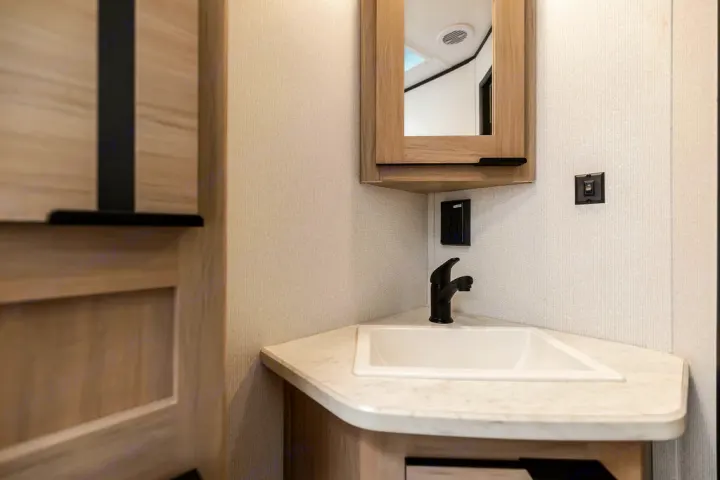 An RV rental's compact bathroom features a corner sink, black faucet, and light wood vanity with a mirrored medicine cabinet.. North Trail 24bhs 2021