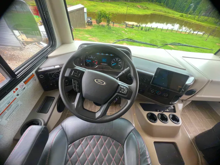 RV rental cockpit view: Ford steering wheel, driver's seat, dashboard, and a scenic pond vista through the windshield.. Thor Outlaw 2022