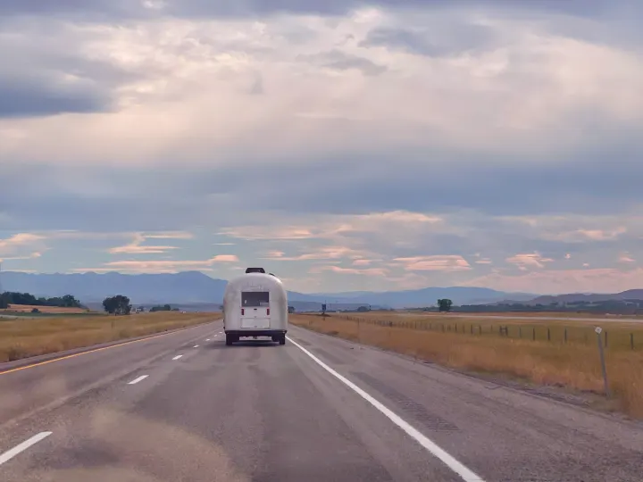 Silver Airstream RV rental towed on highway with mountains & cloudy sky. Perfect for scenic road trips.. Airstream Safari 1964
