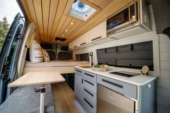This RV rental showcases a modern camper van interior with a cozy bed, well-equipped kitchen, and a dining table.. Mercedes Sprinter 2021