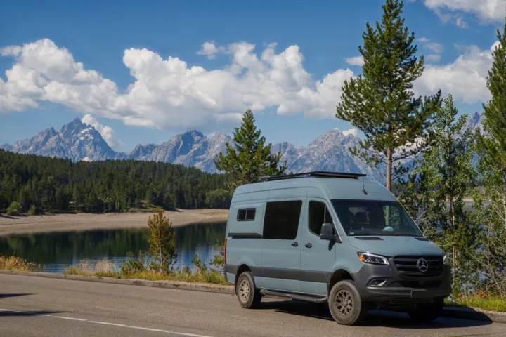 This light blue RV rental Sprinter van is parked by a scenic lake with majestic mountains under a blue, cloudy sky.. Mercedes Sprinter 2021