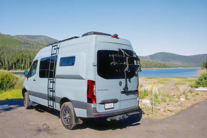 Stylish grey Mercedes Sprinter RV rental parked by a serene lake and forest, featuring a ladder and bike rack.. Mercedes Sprinter 2021