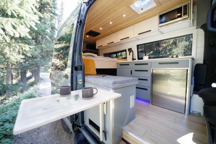 Explore this modern rv rental's interior: wood ceiling, kitchen, bed, and a pull-out table with mugs outdoors.. Mercedes Sprinter 2021