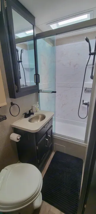 Inside a modern RV rental bathroom. Features a compact shower-tub, toilet, and vanity with dark wood cabinets and a light counter.. Jayco Greyhawk 2021