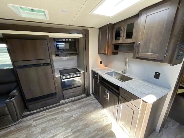 RV rental kitchen interior: modern dark wood cabinets, stainless sink, stove, microwave, and fridge.. Grand Design Transcend Xplor 2022