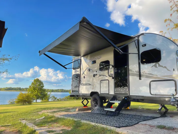 Experience nature with this RV rental! A travel trailer set up lakeside, featuring an extended awning and outdoor mat.. Forest River Cherokee Wolf Pup Black Label 2022