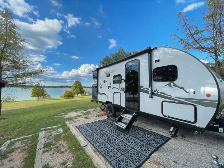 Serene RV rental setup: white travel trailer parked by a lake with an outdoor mat on the gravel site.. Forest River Cherokee Wolf Pup Black Label 2022