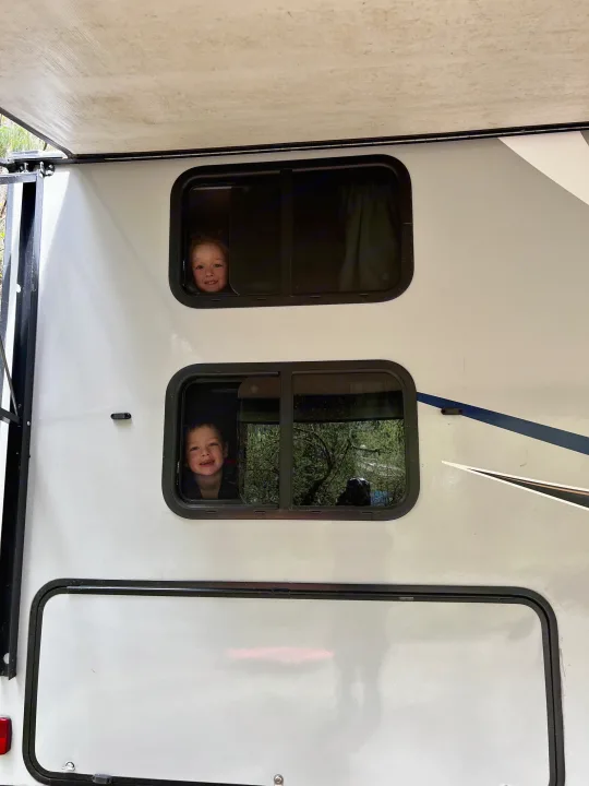 Smiling kids peer from an rv rental's side windows. A white camper with an awning, surrounded by nature.. Coachmen Apex 2018