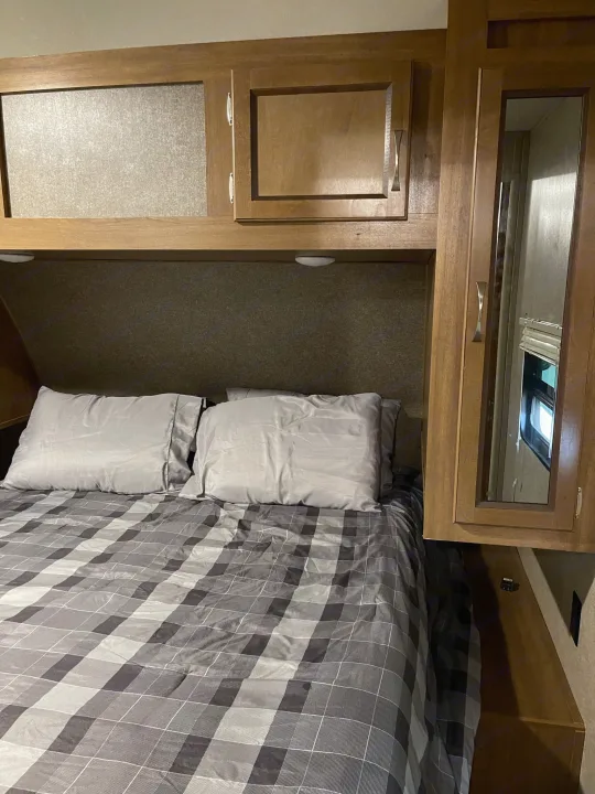 Cozy RV rental bedroom featuring a bed with gray plaid bedding, overhead cabinets, and a mirrored closet door.. Coachmen Catalina 2019