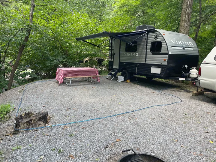 RV rental on a gravel campsite, featuring an extended awning, picnic table, and surrounded by lush trees.. Other 1906st Saga 2021