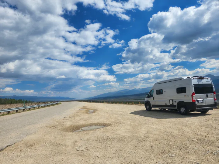 Silver RV rental parked at a scenic mountain overlook. Winding road, distant peaks, and blue sky with white clouds.. Winnebago Solis 2022