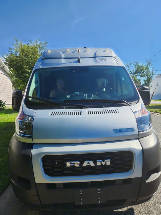 Front view of a light Winnebago Ram van, ideal for RV rental, with a dog "driving" and a person visible inside.. Winnebago Solis 2022