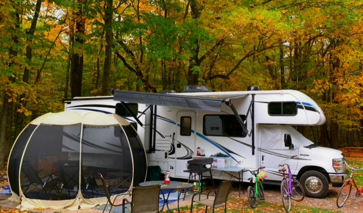 Enjoy fall camping with an RV rental! White RV set up at a forest campsite, featuring a screen tent, bikes, and outdoor gear.. Forest River Sunseeker LE 2020