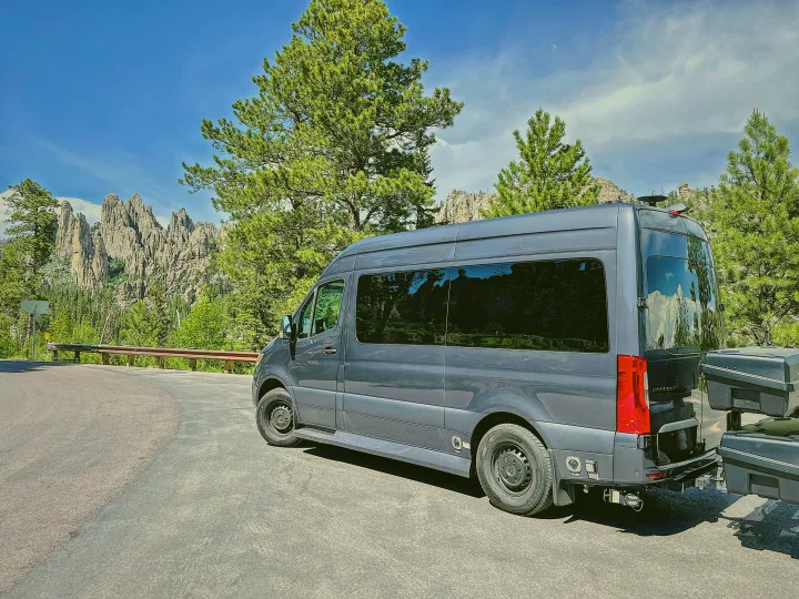 A gray Mercedes Sprinter rv rental van pauses on a scenic mountain road, surrounded by jagged rock formations and pine trees.. Ultimate Toys Ultimate Cruiser 2022