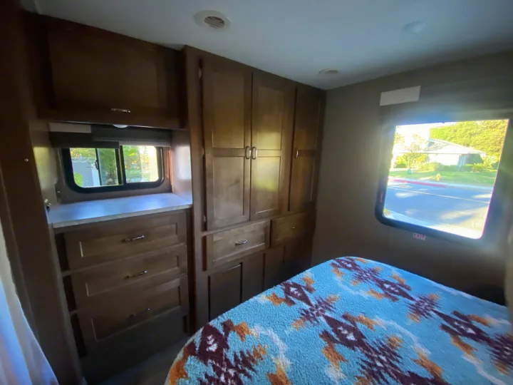 Cozy RV rental bedroom with ample storage and bright windows, featuring a patterned blanket and wood cabinetry.. Winnebago Outlook 2019