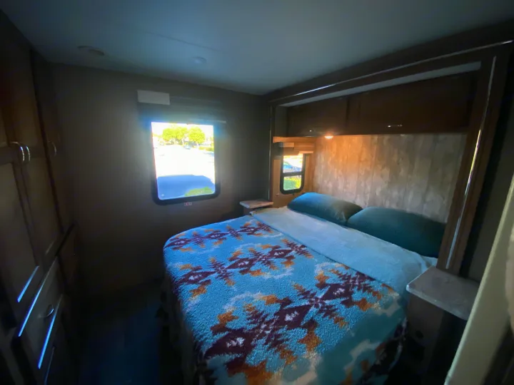 Cozy RV rental bedroom with a patterned bed, pillows, and bright windows, perfect for travel.. Winnebago Outlook 2019