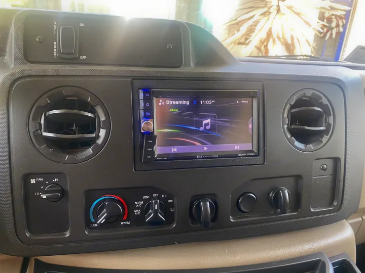 Dashboard of an RV rental featuring a streaming car stereo, climate controls, air vents, and a battery boost switch.. Winnebago Outlook 2019