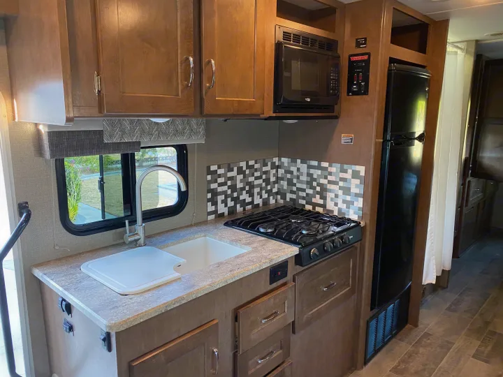 RV rental kitchen with wood cabinets, sink, stove, microwave, and fridge, bathed in natural light from the window.. Winnebago Outlook 2019