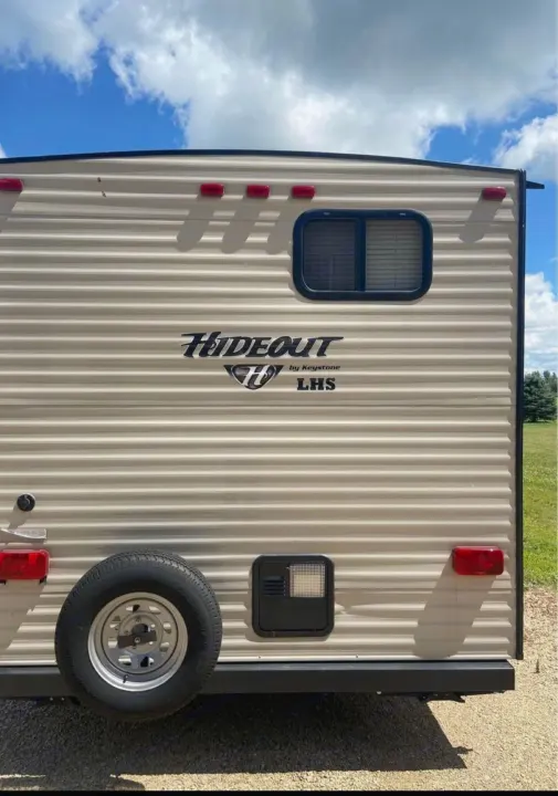 Rear view of a beige rv rental, showing its spare tire, back window, and "Hideout" decal under a blue sky.. Keystone Hideout 2016