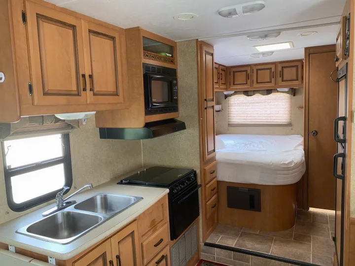 Cozy rv rental interior with a bright kitchen featuring a double sink, stove, microwave, and wood cabinets, next to a bedroom with a white bed.. Forester Forester Motorhome 2010
