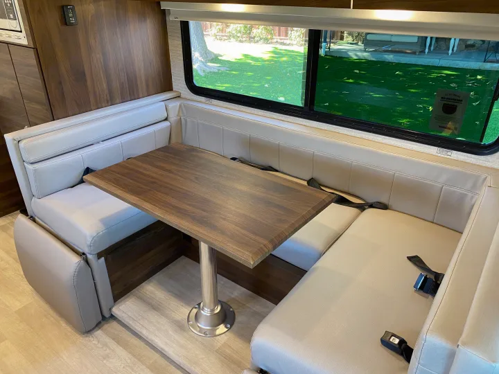 RV rental interior showing a cozy dinette area with light upholstered benches, a wooden table, and a window view of greenery.. Winnebago View 2022