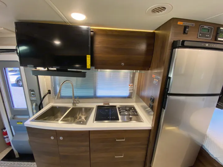 RV rental interior: modern kitchen with dual sink, stovetop, fridge, mounted TV, and controls. A bed is also visible.. Winnebago View 2022